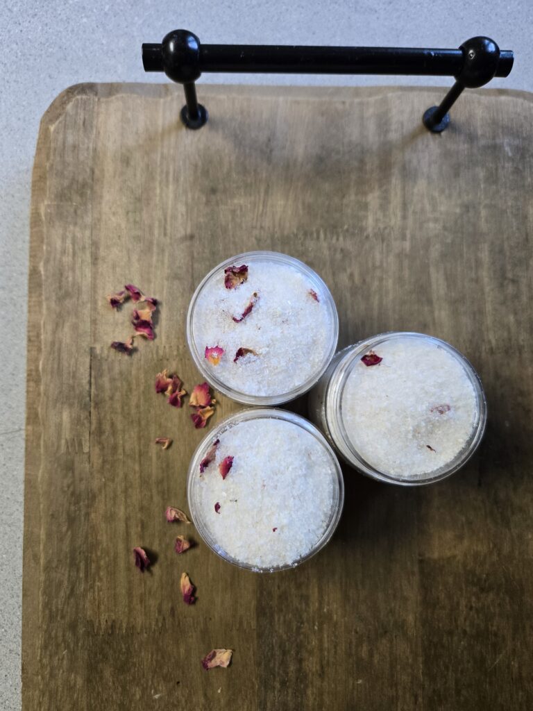 Three bath soaks sitting on a wooden tray surrounded by rose petals