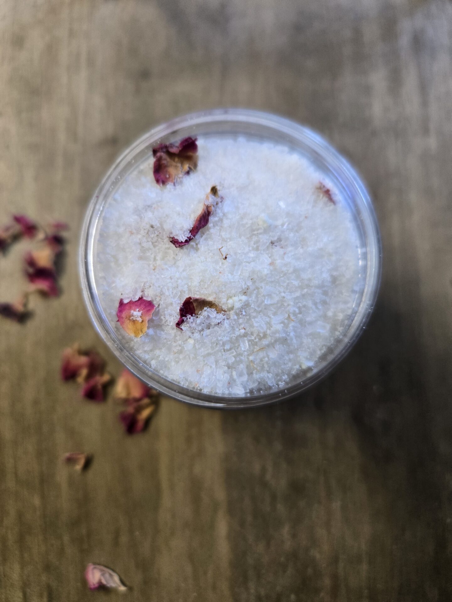 A jar of homemade bath soak on a wooden board surrounded by rose petals.